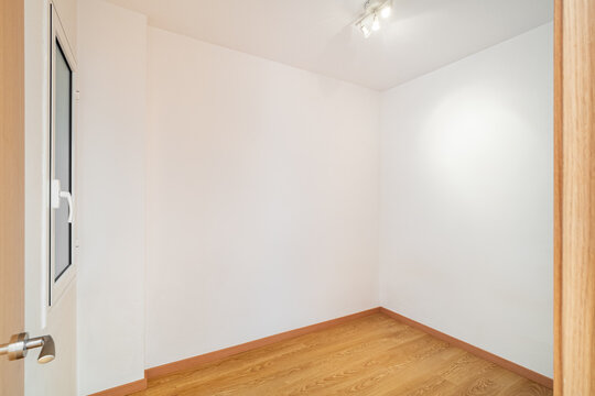 Empty room with white walls and wooden floor. A door is visible on the left side. The space is well-lit with a ceiling light.
