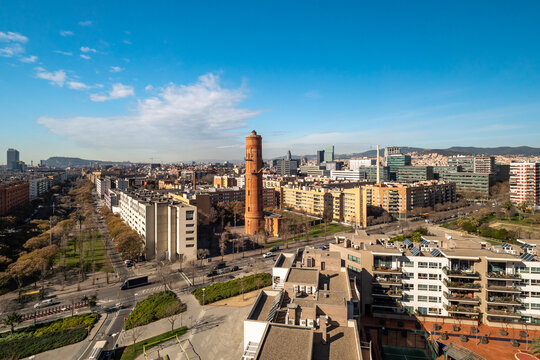 Aerial view of Barcelona showcasing urban architecture, including residential buildings and the iconic red brick tower. Clear blue sky enhances the vibrant cityscape.