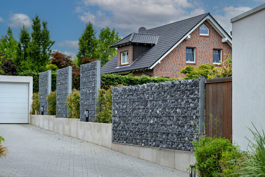 A stylish gabion stone wall and wire mesh fence standing next to a modern paved driveway of a residential brick house.