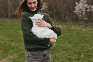 Obraz premium Lifestyle outdoor scene with a woman in a knitted sweater holding a white goat kid in a grassy park, calm playful mood, trees in bloom, natural rustic setting and a gentle companionship in nature.