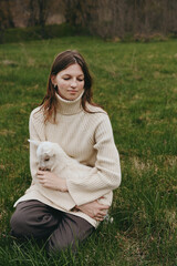 Obraz premium Calm woman outdoors, seated on grass, holding a goat kid, cozy cream sweater, soft natural light, tranquil expression, nature background, relaxed lifestyle moment, tender connection