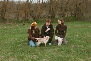 Obraz premium Three friends outdoors in a green field interact with two young goats, smiling and petting them. A warm, candid lifestyle moment amid countryside scenery and natural light, capturing companionship