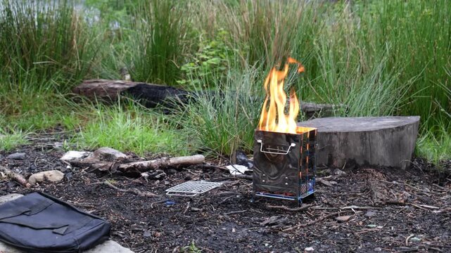 Foldable camp stove with large flames burns on forest floor at rustic campsite. Static shot shows portable metal firebox, kindling area, and summer greenery outdoors.
