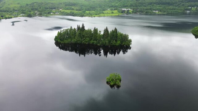 Drone orbit of Eilean Gorm, Loch Ard in Scotland, circling the tree-covered island above dark reflective water with hills and shoreline in the distance.