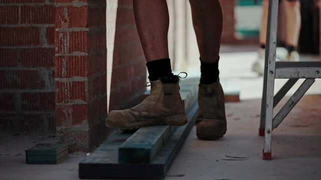Close-up of construction worker wearing PPE safety boots and socks while walking along timber formwork inside a building under construction. Emphasizes workplace safety and protective footwear.