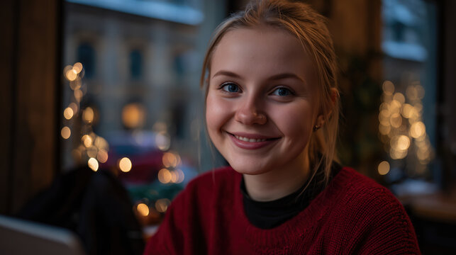 Smiling young woman in red sweater using laptop inside cozy cafe during evening with warm lights suggesting friendly remote work study lifestyle social media learning comfort personal growth
