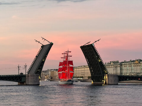 Illuminated tall ship with beautiful red sails sailing under an open bascule bridge on a wide river during a vibrant sunset in a historic city.