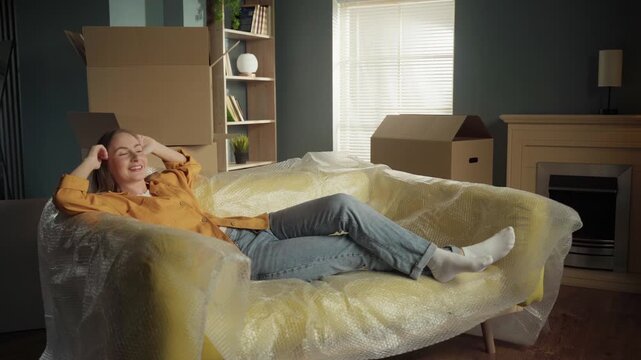 Young happy woman joyfully jumping backwards onto a new sofa covered in bubble wrap, relaxing and celebrating moving into her new home surrounded by cardboard boxes in the living room
