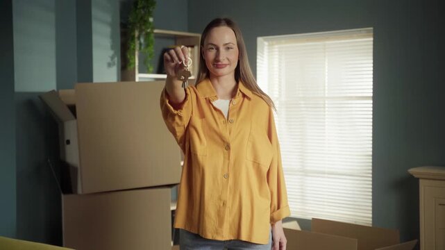 Happy young woman standing in a new apartment with cardboard boxes, smiling and proudly showing the keys to the camera after successfully moving in. Real estate and home ownership concept