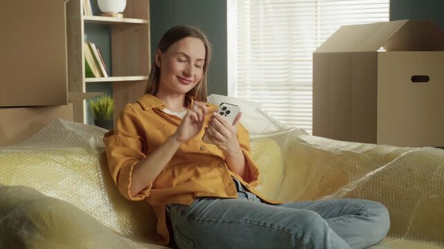 Smiling young woman sitting on a bubble wrapped sofa in her new apartment, happily using her smartphone to browse online amidst cardboard boxes, enjoying a moment of relaxation during moving day