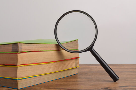 Stack of books with magnifying glass on wooden table, representing research, education, knowledge acquisition, academic study, learning, and information discovery.