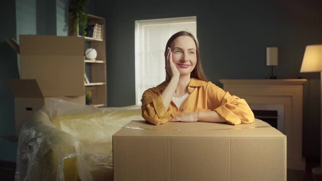 Contented young woman smiling and daydreaming while leaning on a cardboard box in her new apartment, surrounded by packed furniture and looking forward to a fresh start