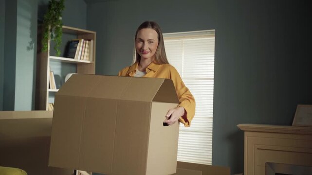 Smiling caucasian woman carrying a heavy cardboard box during the moving process in her new modern apartment, starting a new chapter of life with excitement and joy as a first time homeowner