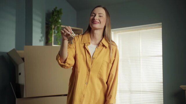 Joyful young woman excitedly holding the keys to her new apartment, looking around the room filled with cardboard boxes, and celebrating the beginning of a new chapter in her life