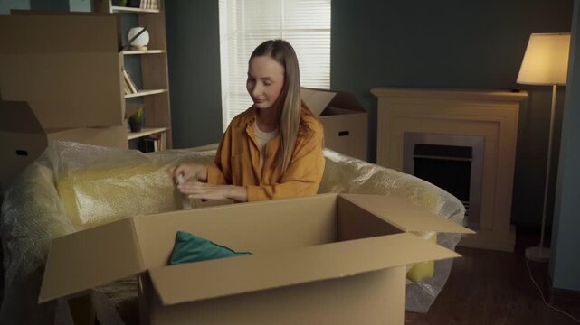 Caucasian woman carefully packing her personal belongings like pillows, photo frames, and books into a large cardboard box in her living room, preparing for the big move to a new apartment