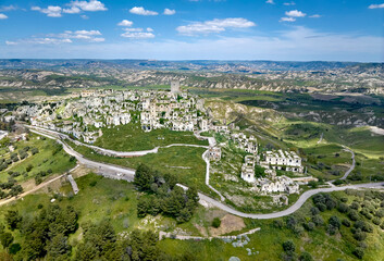 Obraz premium Aerial drone view of the abandoned ghost village of Craco in Basilicata, Italy, showing dramatic hilltop ruins, winding road, green valleys and rugged cliffs under a clear blue sky