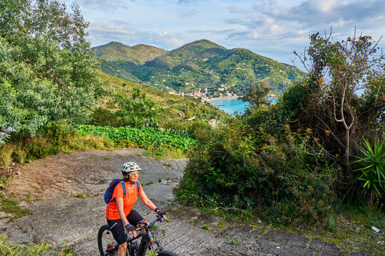 Active senior woman riding an e-bike on a scenic coastal road with panoramic sea and mountain views under dramatic skies. Levanto, Cinque Terre, Liguria, Italy