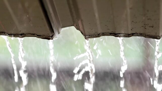 Close up of heavy rainwater flowing from a roof gutter spout during a storm. Pouring water stream with a beautifully blurred green garden bokeh background texture.