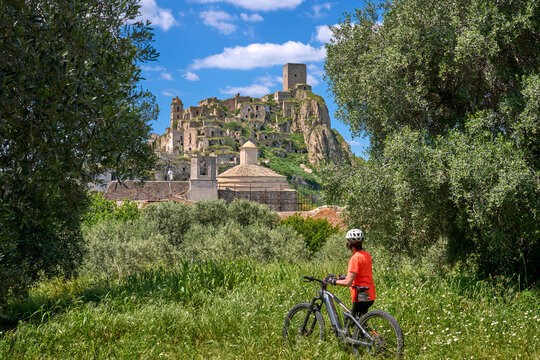 Senior woman cycling an electric mountain bike on a rural road below the abandoned ghost village of Craco in Basilicata, Italy. Dramatic cliff landscape, historic ruins, green fields and blue sky 