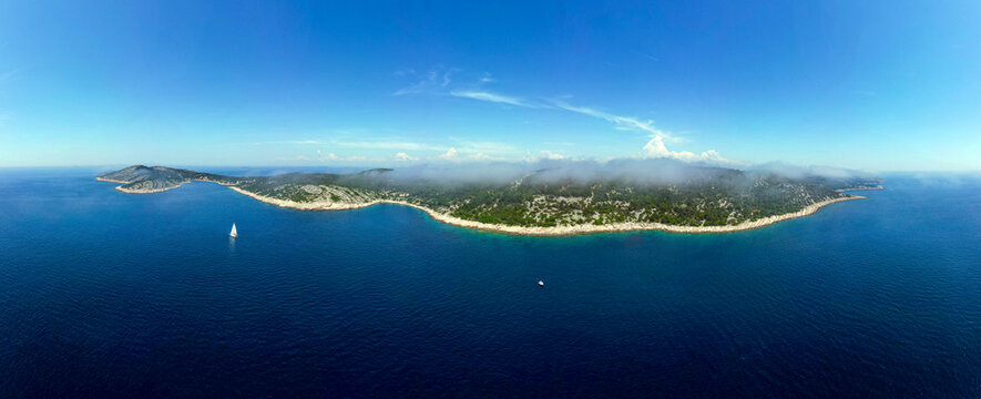 Aerial view of the rocky coastline of the island with green vegetation and a white sailboat on the blue Adriatic Sea in Zirje, Sibensko-kninska zupa, Croatia.