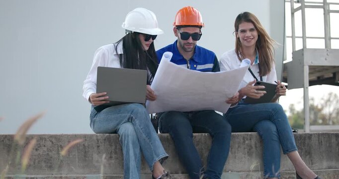 A team of three engineers in hardhats and safety vests review blueprints and discuss a renewable energy project while inspecting wind turbines and planning wind farm development outdoors.
