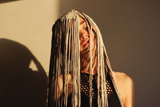 woman with artistic hair styled with many hanging threads covering her face, illuminated by a warm light.