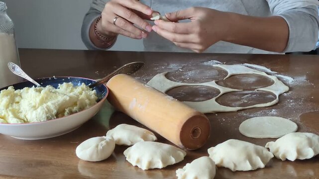 Close-up of a woman's hands expertly shaping traditional Polish pierogi dumplings on a flour-dusted wooden kitchen table, featuring a rolling pin, raw dough, and fresh potato cheese filling.