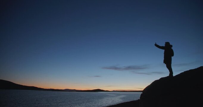 Silhouette of a man standing on a rocky shore and taking a selfie with a smartphone at twilight. Dark figure against a deep blue evening sky and calm water creates a dramatic outdoor travel scene with