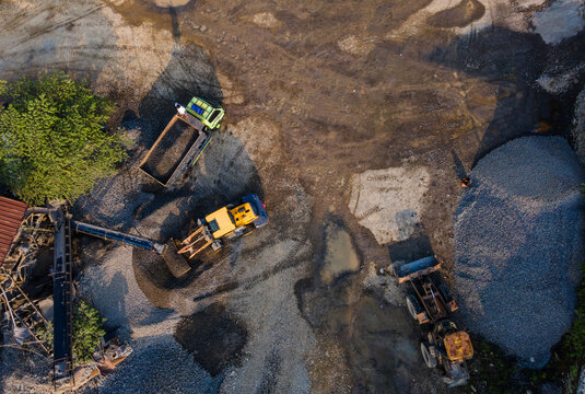 Aerial view of heavy machinery including excavators and trucks working at a gravel quarry site with piles of stones in Karanglewas, Central Java, Indonesia.