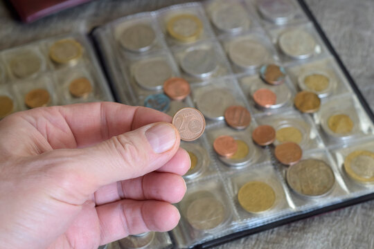 Man holds two euro cent coin against blurred background of numismatic albums designed to store coin collection. Close-up.