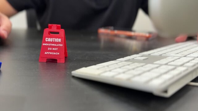 Close-up shot of a small red caution sign reading overstimulated do not approach on a black desk. A person's blurred hands type on a white computer keyboard in the bright indoor background.
