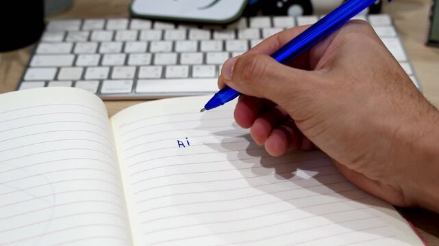 Close-up shot of a person's hand using a blue pen to write AI content planning notes in a lined notebook. The indoor desk workspace features a blurred computer keyboard in the background.