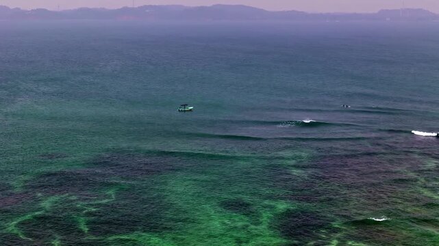 A lone boat and a few surfers sit beyond shallow green reef as small waves roll across Weligama Bay.