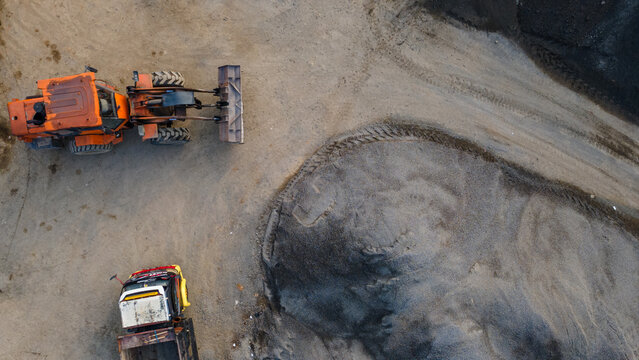 Aerial view of an orange wheel loader and a truck working at a construction site with piles of gravel and dirt tracks in Karanglewas, Central Java, Indonesia.