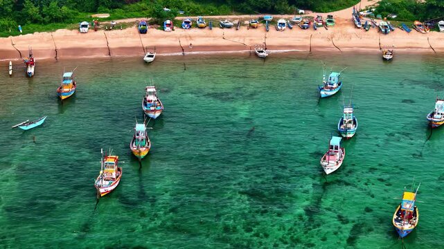Colorful wooden fishing boats float in calm clear green water off a sandy beach lined with parked craft and trees.