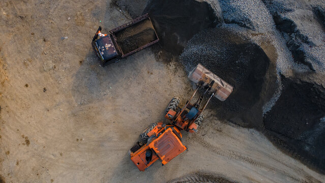 Aerial view of an orange wheel loader and a truck working at a gravel quarry with large piles of dark stone in Karanglewas, Central Java, Indonesia.
