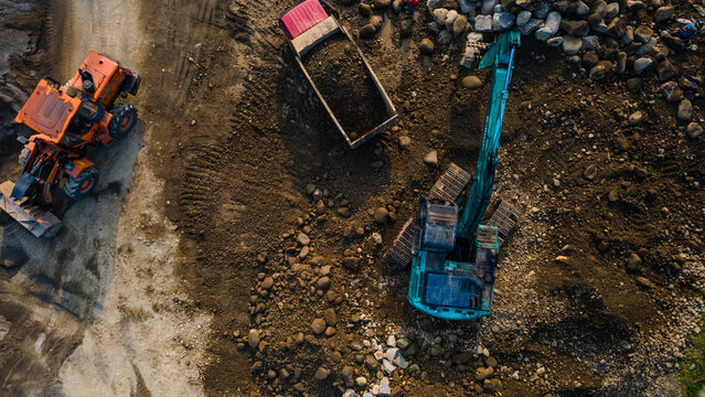 Aerial view of a turquoise excavator loading a dump truck next to an orange wheel loader at a construction site with piles of rocks in Karanglewas, Central Java, Indonesia.