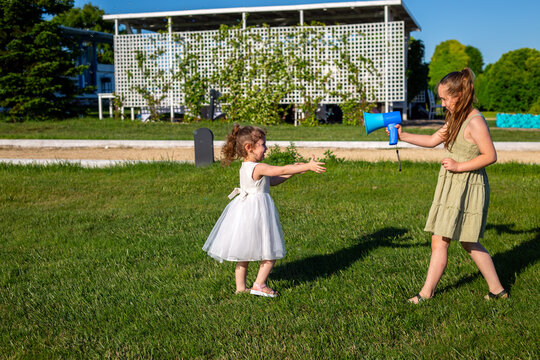 Two girls playing with blue bubble gun on green lawn near white lattice fence with soap bubbles in sunny park