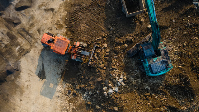 Aerial view of a teal excavator and an orange wheel loader moving large rocks and soil at a construction site in Karanglewas, Central Java, Indonesia.