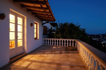 Obraz premium Balcony view at twilight with illuminated house windows, balustrade, and distant city lights. Peaceful outdoor space with plants, featuring Mediterranean architecture and warm lighting on tiled floor.