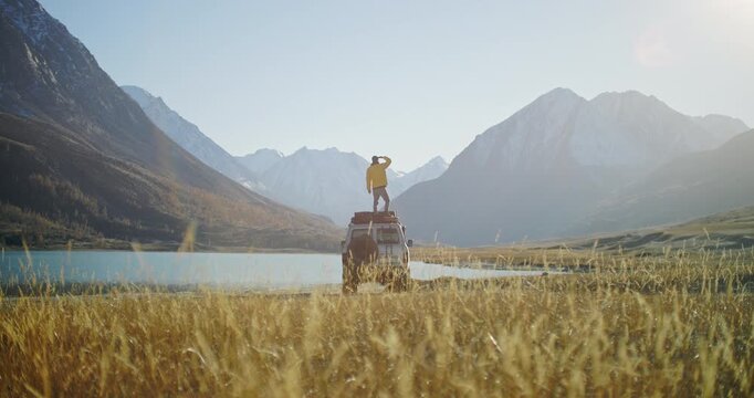 Traveler in a yellow jacket standing on the roof of an off road SUV and looking across a mountain lake toward snowy peaks and a wide alpine valley. Golden grass, bright daylight, and remote wilderness