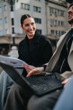 Two colleagues attend a winter business meeting outdoors, reviewing documents and a laptop while smiling. A professional pair converse and share ideas on a bench during an outdoor work discussion.