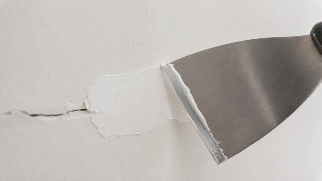 A stainless steel putty knife smoothing white spackle over a wall crack.