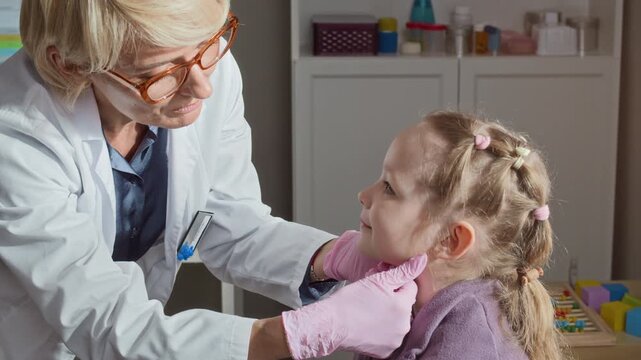 Caucasian middle aged female pediatrician examining lymph nodes of little girl in clinic. Clip is perfect for pediatric healthcare marketing and medical clinic promotional videos