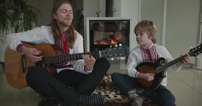 Man and Kid Boy Play Music on Guitar and Mandolin Sitting on Floor Near Fireplace. 4K 10-bit