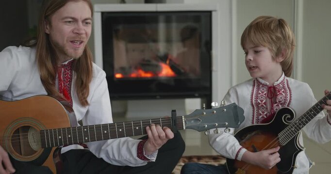 Man and Kid Boy Play Music on Guitar and Mandolin Sitting on Floor Near Fireplace. 4K 10-bit