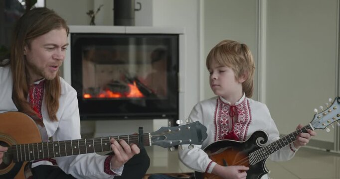 Man and Kid Boy Play Music on Guitar and Mandolin Sitting on Floor Near Fireplace. 4K 10-bit