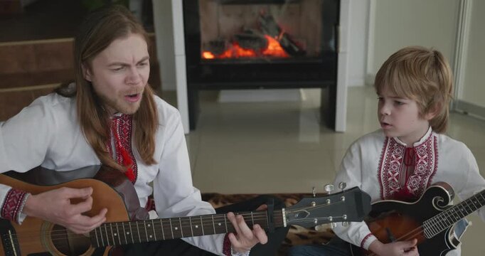 Man and Kid Boy Play Music on Guitar and Mandolin Sitting on Floor Near Fireplace. 4K 10-bit