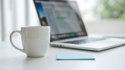 Fototapeta premium A white ceramic coffee mug on a desk next to an open silver laptop computer
