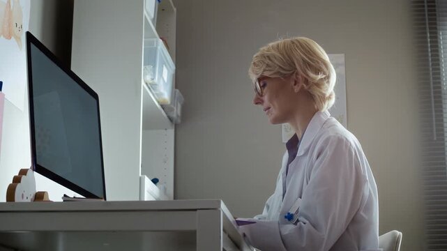 Low angle shot of middle aged Caucasian female pediatrician sitting at desk and typing on computer in clinic office. Clip is suitable for healthcare software and medical clinic marketing campaigns